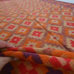 Op de afbeelding: Een close-up van een kleurrijk geweven tapijt met een geometrisch patroon in tinten oranje, rood, paars en beige. Het tapijt is dubbelgevouwen, waardoor de ingewikkelde details van de weving zichtbaar worden.