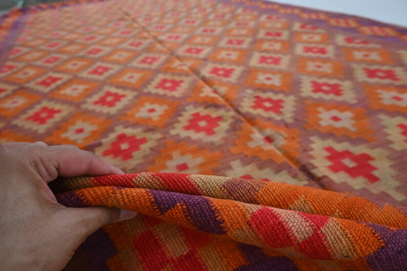 Op de afbeelding: Een close-up van een kleurrijk geweven tapijt met een geometrisch patroon in tinten oranje, rood, paars en beige. Het tapijt is dubbelgevouwen, waardoor de ingewikkelde details van de weving zichtbaar worden.
