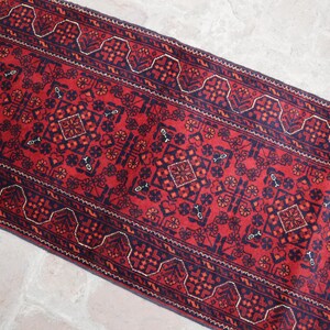 May include: A rectangular, red runner rug with a detailed geometric pattern. The rug features a central design with diamond and floral motifs, framed by a dark blue border with repeating shapes. The rug's design is symmetrical and traditional.