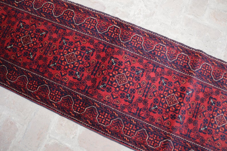 May include: A rectangular, red runner rug with a detailed geometric pattern. The rug features a central design with diamond and floral motifs, framed by a dark blue border with repeating shapes. The rug's design is symmetrical and traditional.