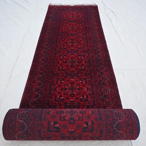 Puede incluir: Una alfombra pasillera larga y rectangular con una base de color rojo intenso y patrones geométricos y florales intrincados en negro y azul oscuro. La alfombra está parcialmente enrollada en un extremo, revelando un diseño denso y simétrico.