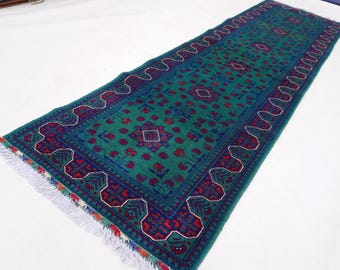 May include: A long, rectangular rug with a teal field and a navy blue border. The rug features a repeating pattern of red and navy blue geometric shapes and a white fringe on both ends. The rug is likely made of wool.