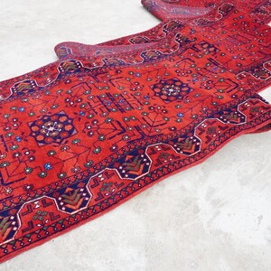 May include: A red runner rug with a traditional Persian design featuring intricate blue and green patterns. The rug is folded in half, showcasing the detailed craftsmanship.