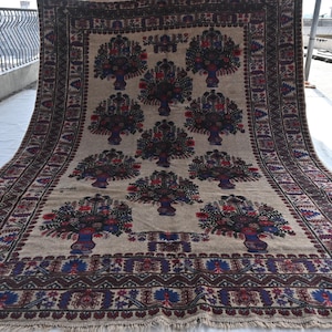 Può includere: Un tappeto rettangolare beige con un motivo ripetuto di composizioni floreali in vasi. I fiori sono rossi, blu e viola. Il tappeto ha un bordo scuro con disegni geometrici e frange.
