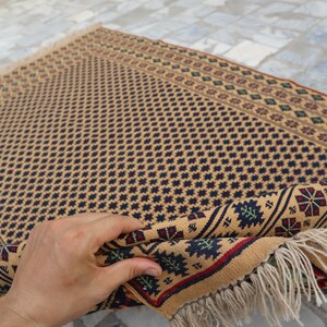 May include: A beige rug with a geometric pattern in shades of blue, red, and green. The rug has a fringe border and is folded in half.