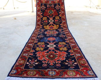 May include: A long, narrow rug with a navy blue background and intricate floral patterns in shades of red, yellow, and blue. The rug has a white fringe on both ends.
