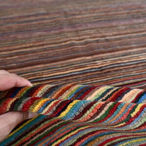 Pode incluir: Um tapete listrado colorido com uma m&atilde;o segurando uma se&ccedil;&atilde;o do tapete para mostrar o detalhe das listras. O tapete &eacute; feito de um material macio e felpudo e tem uma variedade de cores, incluindo vermelho, azul, verde, amarelo e marrom.