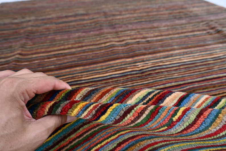 Pode incluir: Um tapete listrado colorido com uma m&atilde;o segurando uma se&ccedil;&atilde;o do tapete para mostrar o detalhe das listras. O tapete &eacute; feito de um material macio e felpudo e tem uma variedade de cores, incluindo vermelho, azul, verde, amarelo e marrom.