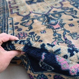 Peut inclure: Une section d'un tapis vintage avec un fond beige et des motifs complexes en bleu marine, rose et bleu clair. La bordure du tapis pr&eacute;sente un motif g&eacute;om&eacute;trique d&eacute;taill&eacute;. Le tapis est soulev&eacute;, r&eacute;v&eacute;lant sa texture.