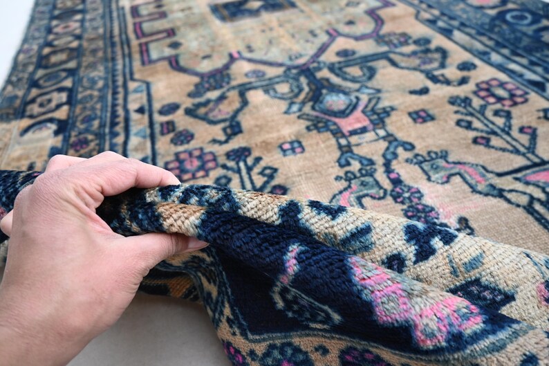 Peut inclure: Une section d'un tapis vintage avec un fond beige et des motifs complexes en bleu marine, rose et bleu clair. La bordure du tapis pr&eacute;sente un motif g&eacute;om&eacute;trique d&eacute;taill&eacute;. Le tapis est soulev&eacute;, r&eacute;v&eacute;lant sa texture.