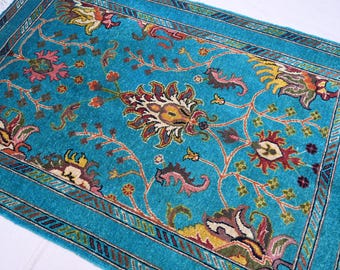 May include: A turquoise blue rug with a floral pattern. The rug has a border with a repeating geometric design. The flowers are in shades of pink, yellow, red, and green.