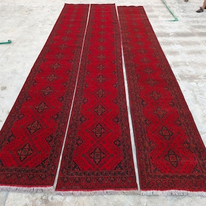 K&ouml;nnte beinhalten: Drei lange, rechteckige rote Teppiche mit komplizierten geometrischen Mustern in Schwarz und Braun. Die Teppiche sind nebeneinander angeordnet und zeigen ihre L&auml;nge und ihr Design. Die Teppiche haben Fransen an den Enden.