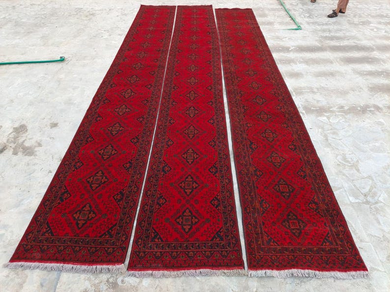 K&ouml;nnte beinhalten: Drei lange, rechteckige rote Teppiche mit komplizierten geometrischen Mustern in Schwarz und Braun. Die Teppiche sind nebeneinander angeordnet und zeigen ihre L&auml;nge und ihr Design. Die Teppiche haben Fransen an den Enden.
