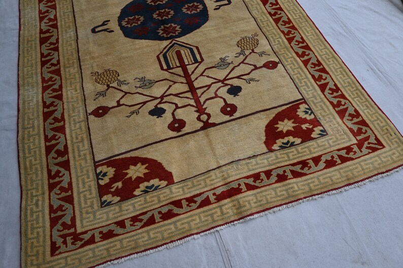 May include: A beige rug with a red, blue, and green border. The center of the rug features a stylized tree with red and blue fruit, and a blue and red circle with stars.