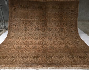 May include: A large rectangular area rug in shades of brown and tan, featuring a repeating geometric pattern of squares and crosses across the center field, enclosed by a wide decorative border with intricate motifs and a short fringe edge.