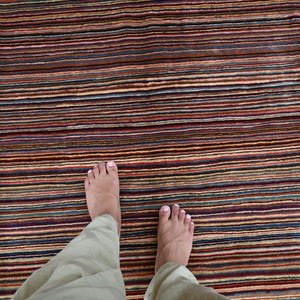 Pode incluir: Os p&eacute;s descal&ccedil;os de uma pessoa est&atilde;o em p&eacute; sobre um tapete listrado multicolorido. O tapete tem uma variedade de cores, incluindo vermelho, azul, verde e marrom.