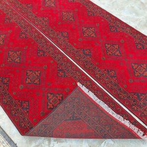 K&ouml;nnte beinhalten: Ein leuchtend roter Teppich mit einem sich wiederholenden geometrischen Muster in Schwarz und Braun. Der rechteckige Teppich hat einen detaillierten Rand und eine Fransenbord&uuml;re. Das Design ist symmetrisch und traditionell.