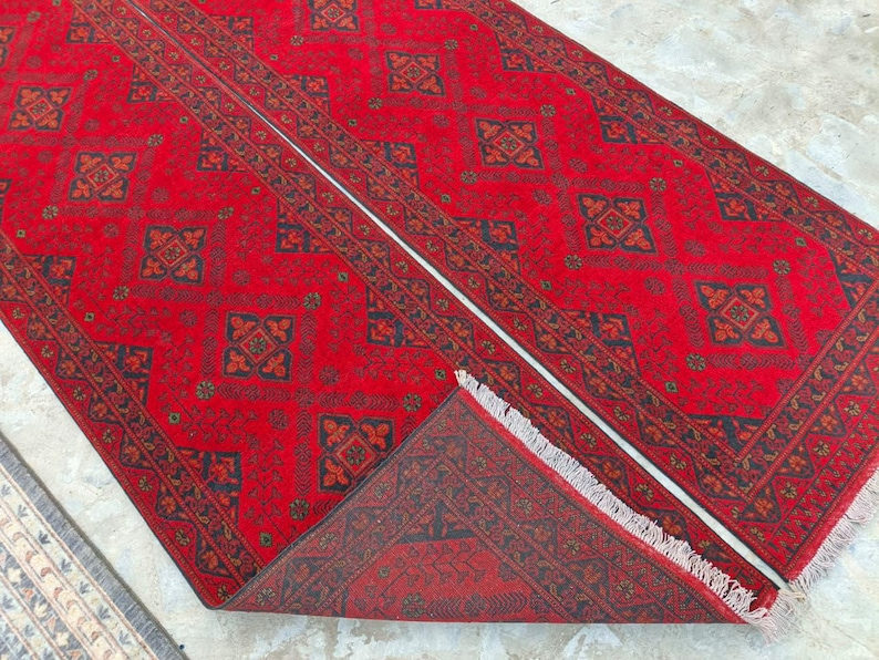 K&ouml;nnte beinhalten: Ein leuchtend roter Teppich mit einem sich wiederholenden geometrischen Muster in Schwarz und Braun. Der rechteckige Teppich hat einen detaillierten Rand und eine Fransenbord&uuml;re. Das Design ist symmetrisch und traditionell.