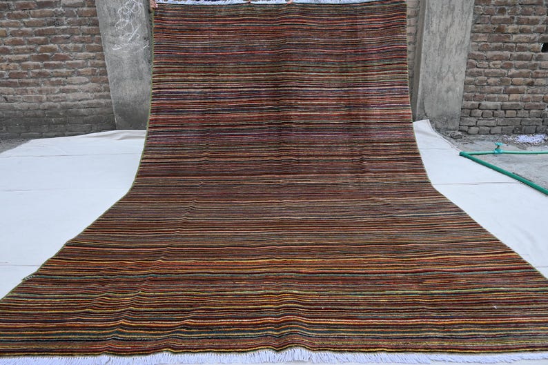 Pode incluir: Um tapete retangular longo com um padr&atilde;o listrado em tons de marrom, vermelho, verde e amarelo. O tapete tem uma borda de franjas brancas.