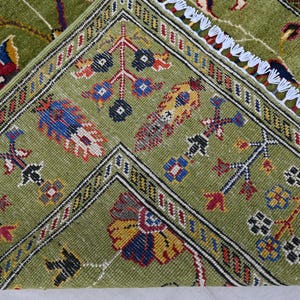 May include: Close-up of a green rug with intricate floral and geometric patterns in shades of red, blue, yellow, and black. The rug has a white fringe border.