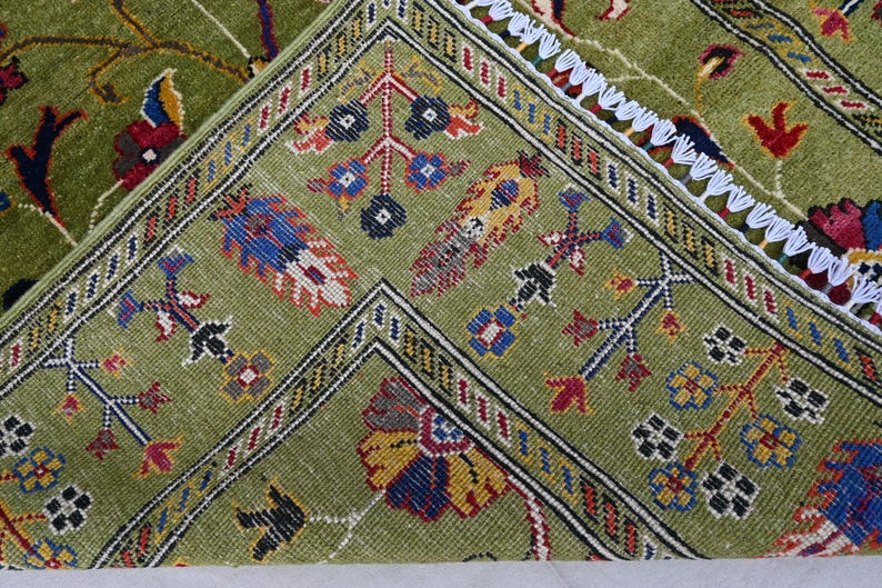 May include: Close-up of a green rug with intricate floral and geometric patterns in shades of red, blue, yellow, and black. The rug has a white fringe border.