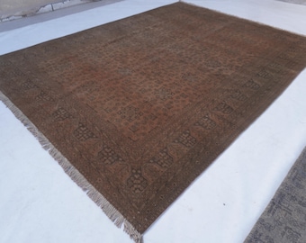 May include: A large rectangular area rug featuring a deep brown and muted bronze colour palette. The rug displays an intricate, repeating geometric pattern across its central field, framed by a wide, detailed border with fringe on the edges.