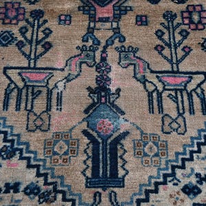 Peut inclure: Gros plan sur un tapis beige avec un motif d&eacute;taill&eacute;. Le motif pr&eacute;sente des cerfs stylis&eacute;s, des arbres et des formes g&eacute;om&eacute;triques dans des tons de bleu et de rose. Le tapis a une bordure avec un motif en zigzag r&eacute;p&eacute;titif.