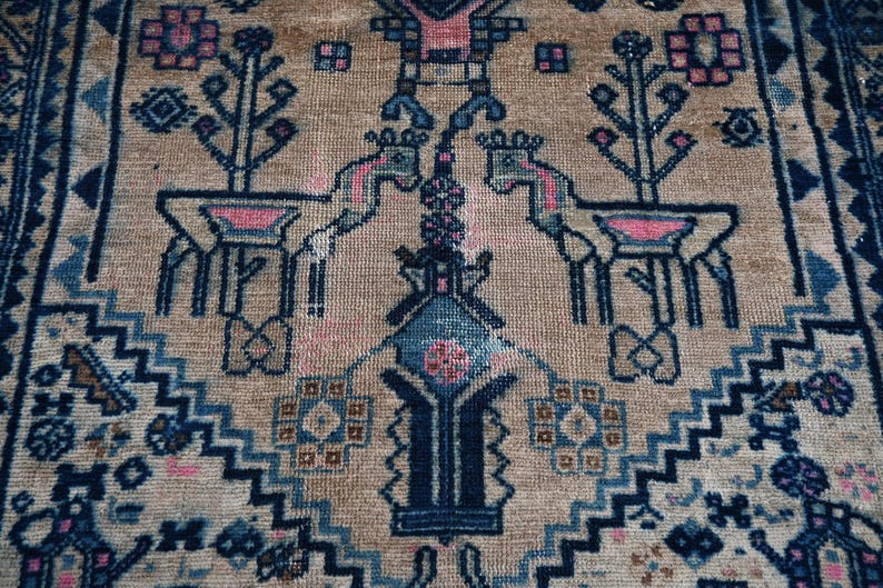 Peut inclure: Gros plan sur un tapis beige avec un motif d&eacute;taill&eacute;. Le motif pr&eacute;sente des cerfs stylis&eacute;s, des arbres et des formes g&eacute;om&eacute;triques dans des tons de bleu et de rose. Le tapis a une bordure avec un motif en zigzag r&eacute;p&eacute;titif.