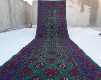 May include: A long, rectangular rug with a green field and a repeating pattern of red and blue floral motifs. The rug has a dark red border with blue and green accents. The rug is displayed on a white surface.