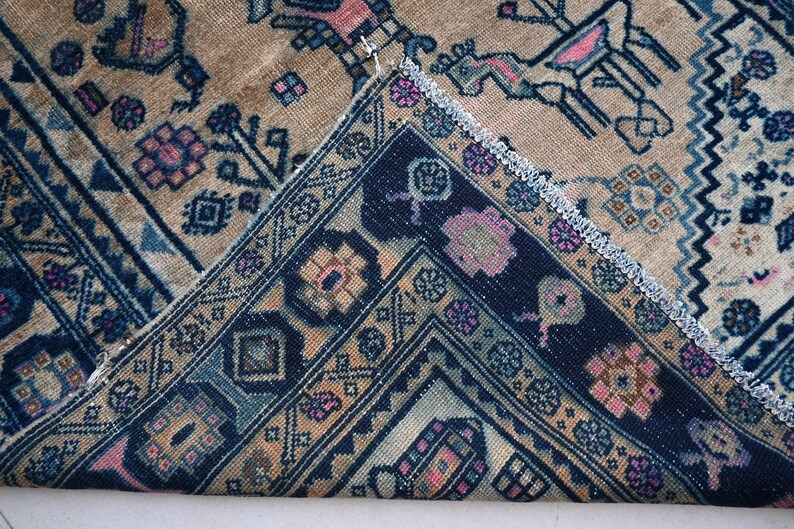 Peut inclure: Un tapis ancien avec un motif g&eacute;om&eacute;trique d&eacute;taill&eacute;. Le tapis pr&eacute;sente un fond beige avec des bordures bleu marine et des accents roses, bleus et bruns. Le design comprend des motifs floraux et animaliers, cr&eacute;ant une esth&eacute;tique vintage.