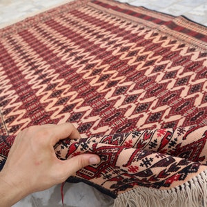 May include: A hand is holding a corner of a red, black and white patterned rug with fringe. The rug has a geometric design.