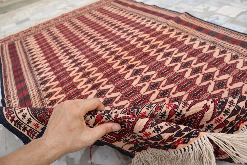 May include: A hand is holding a corner of a red, black and white patterned rug with fringe. The rug has a geometric design.
