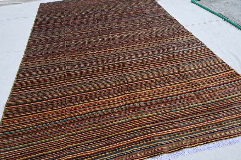 Pode incluir: Um tapete listrado com uma variedade de cores, incluindo marrom, vermelho, laranja, amarelo, verde e azul. O tapete tem um brilho sutil e uma textura macia.