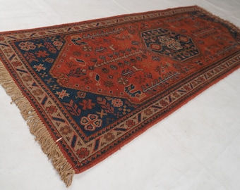 May include: A rectangular area rug featuring a deep orange central field with a dark blue geometric medallion and border. The rug is decorated with intricate floral and geometric motifs in red, blue, and beige, finished with beige fringe.