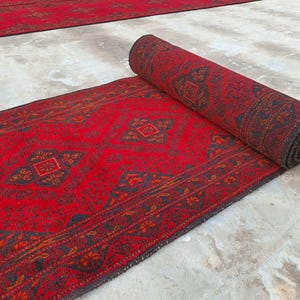 K&ouml;nnte beinhalten: Eine aufgerollte rote Teppichrolle mit detailliertem geometrischem Muster. Der Teppich hat einen dunklen Rand und komplizierte Designs in Rot-, Braun- und Schwarzt&ouml;nen. Der Teppich ist teilweise ausgerollt und zeigt sein volles Muster und seine Textur.