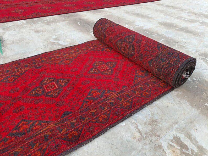 K&ouml;nnte beinhalten: Eine aufgerollte rote Teppichrolle mit detailliertem geometrischem Muster. Der Teppich hat einen dunklen Rand und komplizierte Designs in Rot-, Braun- und Schwarzt&ouml;nen. Der Teppich ist teilweise ausgerollt und zeigt sein volles Muster und seine Textur.