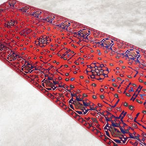 May include: A red runner rug with a traditional Persian design. The rug has a dark blue border with intricate patterns in shades of red, blue, and green.