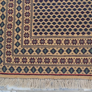 May include: A close-up of a beige rug with a geometric pattern in shades of red, blue, and green. The rug has a fringe border.