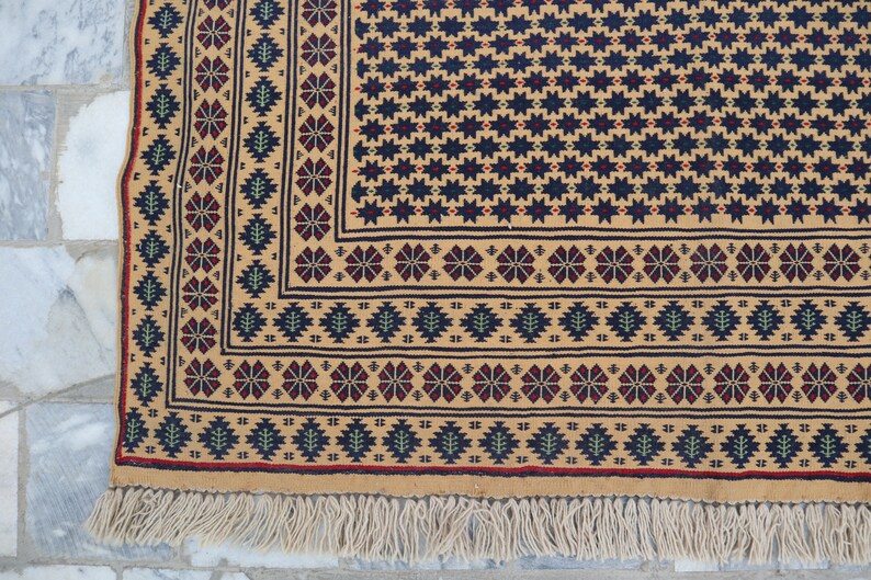 May include: A close-up of a beige rug with a geometric pattern in shades of red, blue, and green. The rug has a fringe border.