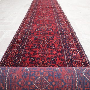 May include: A rolled-up, long, red runner rug with a detailed geometric pattern in navy blue and accents of white. The rug is unrolled on a light-colored stone surface, showcasing its intricate design.