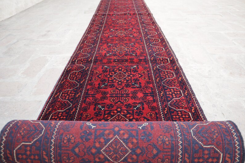 May include: A rolled-up, long, red runner rug with a detailed geometric pattern in navy blue and accents of white. The rug is unrolled on a light-colored stone surface, showcasing its intricate design.