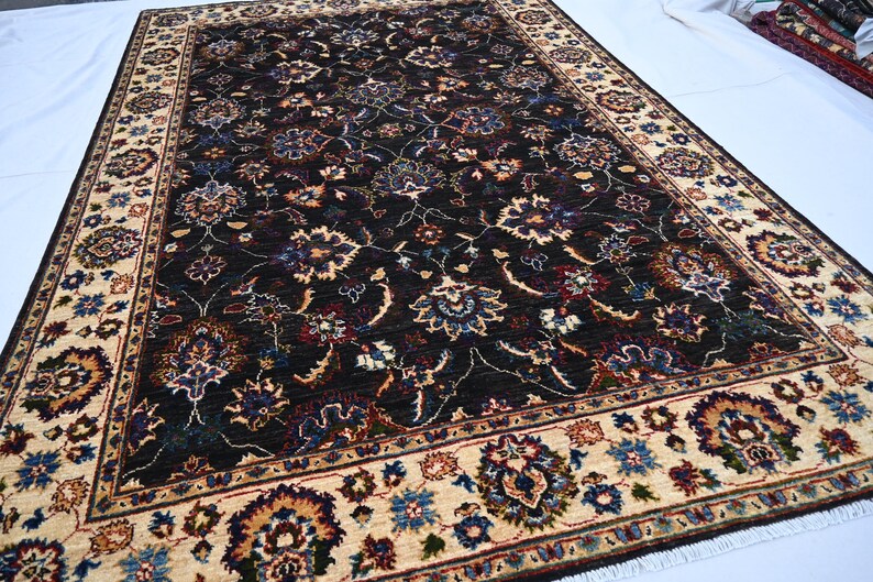 May include: A dark brown rug with a cream-coloured border. The rug has a floral pattern with intricate details in shades of blue, red, and green.