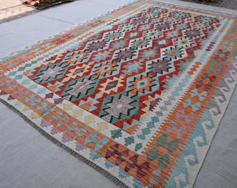 May include: A rectangular, hand-woven rug with a geometric pattern. The rug features a central diamond design in red, orange, and teal, bordered by a cream and orange pattern. The rug's edges have a fringed finish.
