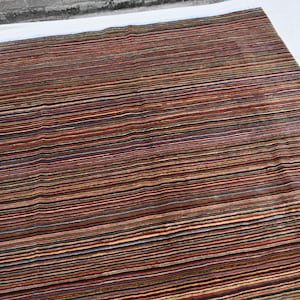 Pode incluir: Um tapete listrado multicolorido com uma variedade de tons de marrom, vermelho, verde e bege. O tapete tem um design contempor&acirc;neo e &eacute; feito de um material macio e felpudo.