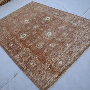 May include: A rectangular, brown rug with a detailed, cream-colored pattern. The rug features a central design with floral motifs and a cream-colored border. The rug is made of a woven material and has a fringe on one edge.