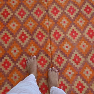Op de afbeelding: De blote voeten van een persoon staan op een kleurrijk tapijt met een geometrisch patroon. Het tapijt is gemaakt van geweven stof en heeft tinten van oranje, rood, paars en cr&egrave;me.