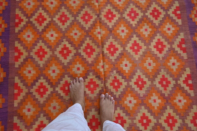 Op de afbeelding: De blote voeten van een persoon staan op een kleurrijk tapijt met een geometrisch patroon. Het tapijt is gemaakt van geweven stof en heeft tinten van oranje, rood, paars en cr&egrave;me.