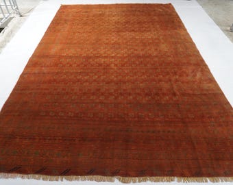 May include: A large rectangular area rug in a deep burnt orange hue, featuring a repeating pattern of small geometric motifs arranged in horizontal bands across the surface, finished with a short, light-colored fringe along the bottom edge.