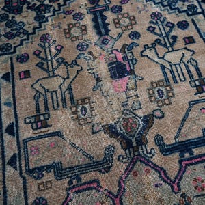 May include: A vintage rug with a beige background and intricate blue and pink patterns. The rug features a repeating design of stylized trees and animals.