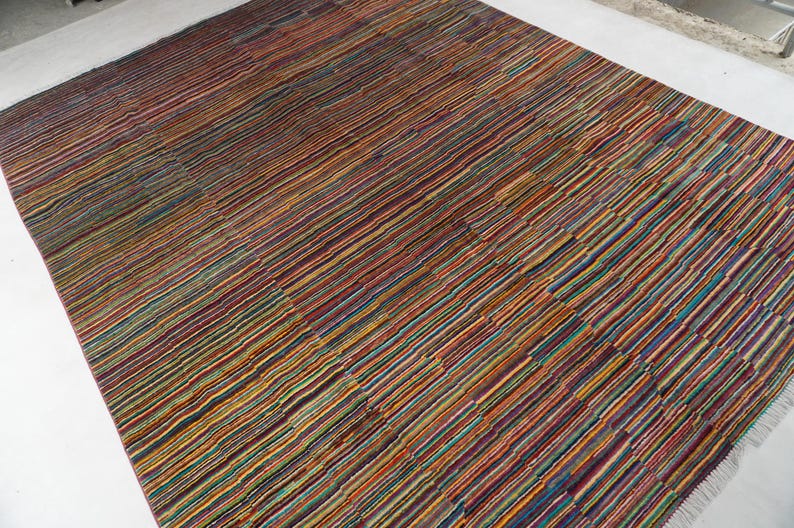 May include: A rectangular area rug featuring a dense, multi-colored striped pattern. The design consists of thin, horizontal lines in various shades of red, orange, yellow, green, blue, and purple, creating a vibrant, textured appearance across the surface.